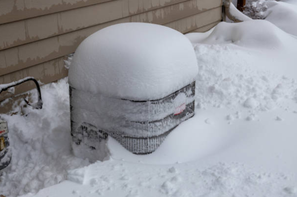 An outdoor HVAC condenser unit is heavily covered in snow, showing the impact of winter weather on home heating and cooling equipment.