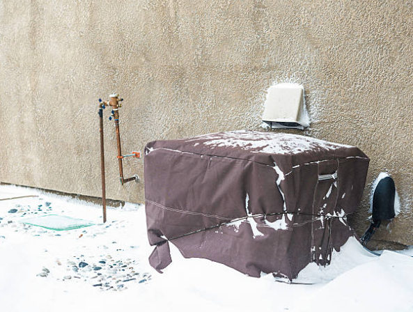 An outdoor air conditioning unit covered with a protective cover is partially buried in snow against a house wall during winter.