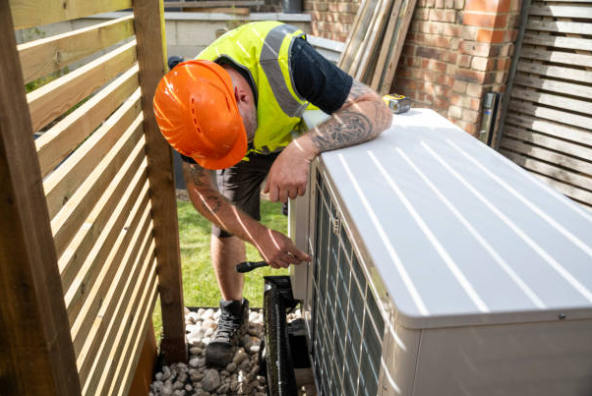 An HVAC technician wearing a hard hat and safety vest services an outdoor air conditioning unit beside a wooden fence.
