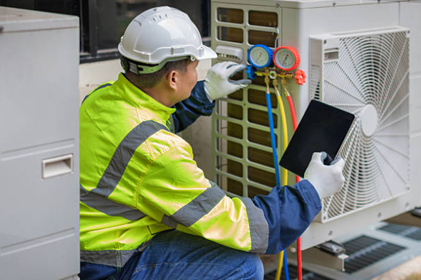 An HVAC technician in safety gear checks refrigerant pressure on an outdoor AC unit using gauges and a tablet for diagnostics.