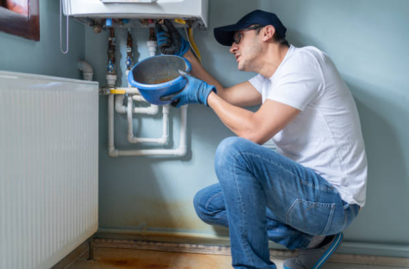 A technician crouches under a wall mounted boiler holding a bucket while checking pipes for a possible water leak.